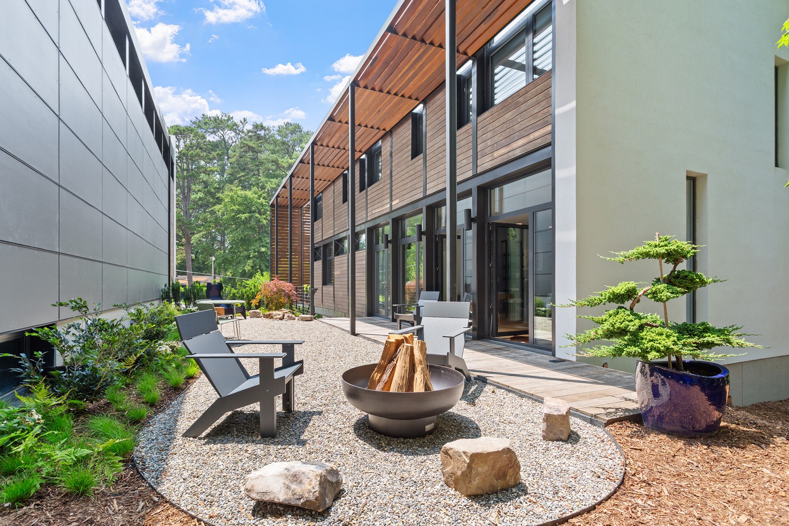 Modern courtyard between contemporary townhomes featuring a gravel seating area with Adirondack chairs, a circular fire pit, landscaped plantings, and expansive floor-to-ceiling windows beneath warm wood soffits.