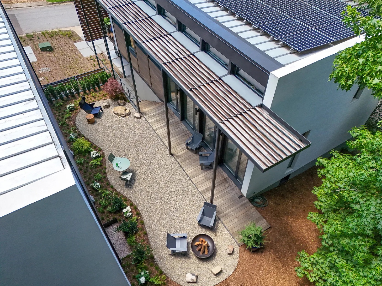 Overhead view of a private landscaped courtyard between modern homes, featuring a spacious wood deck, covered patio, fire pit, gravel seating area, mature landscaping, and rooftop solar panels.